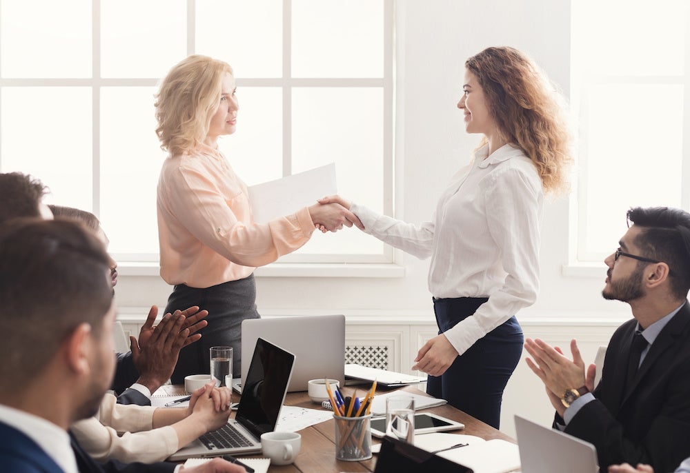 Smiling boss shaking hand of cheerful happy female employee, rewarding excited worker or congratulating partner with achievement, team applauding