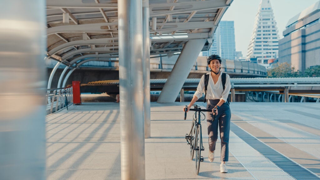 Asian businesswoman go to work at office walk and smile wear backpack look around take bicycle on street around building on a city street. Bike commuting, Commute on bike, Business commuter concept.