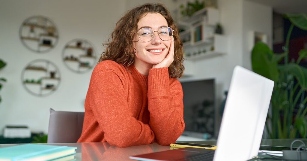 A woman with glasses and a red shirt smiling at the camera while she is in front of her laptop.